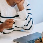 Mobile Education - Positive young African American female freelancer in casual clothes talking on smartphone and drinking coffee while working remotely on laptop at home