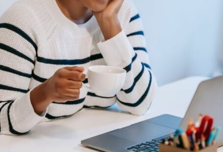 Mobile Education - Positive young African American female freelancer in casual clothes talking on smartphone and drinking coffee while working remotely on laptop at home