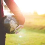 Global Citizenship - person holding black and brown globe ball while standing on grass land golden hour photography