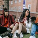 Online University - Full body of happy diverse students with notebooks and laptop sitting on grassy lawn on campus of university while studying together