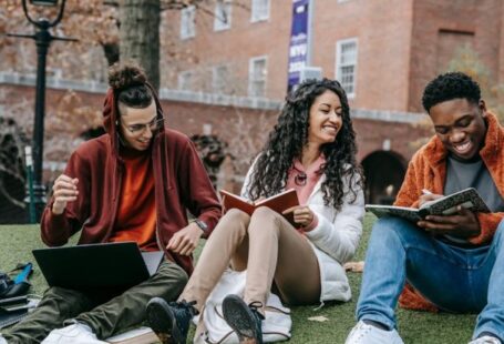 Online University - Full body of happy diverse students with notebooks and laptop sitting on grassy lawn on campus of university while studying together
