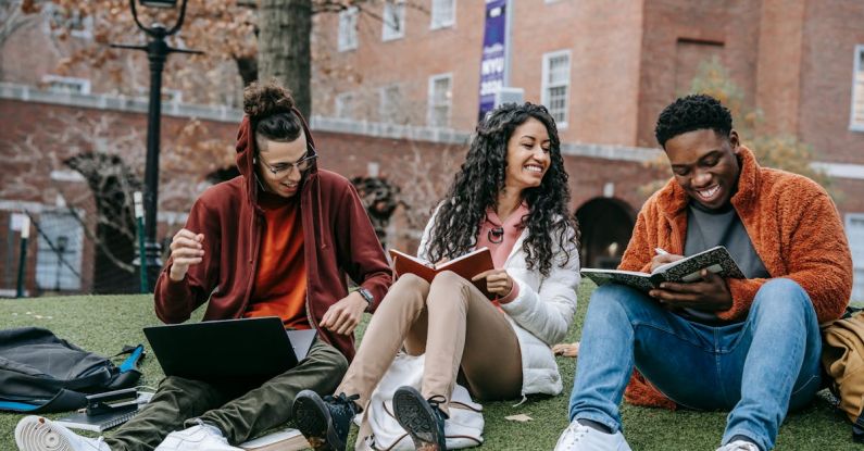 Online University - Full body of happy diverse students with notebooks and laptop sitting on grassy lawn on campus of university while studying together